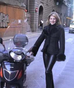 a woman in black leather pants and boots walking next to a motorcycle